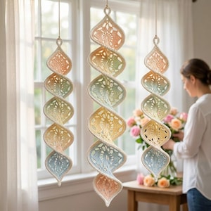 May include: Three hanging spiral ornaments crafted from cream-colored yarn with a delicate openwork crochet pattern. Each ornament features a soft gradient of pastel colors, transitioning from peach and light green to yellow and pale blue.