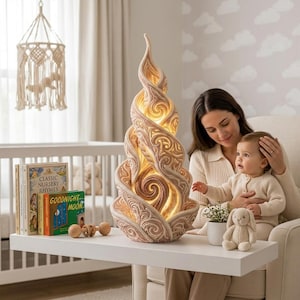 Op de afbeelding: Een hoge, spiraalvormige, crèmekleurige decoratieve lamp met ingewikkelde, uitgesneden wervelpatronen en een warm intern licht. De lamp staat op een wit oppervlak naast een kleine plant in een pot, een pluche konijn en kinderboeken.