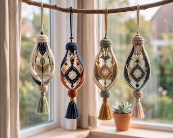 Puede incluir: Cuatro adornos de macramé colgados de una rama de madera. Cada pieza presenta intrincados patrones de nudos, cuentas de madera y una borla inferior. Los adornos muestran variadas paletas de colores que incluyen crema, azul marino, verde salvia y mostaza.