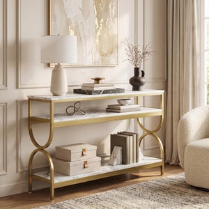 May include: A three-tier console table featuring a gold metal frame with arched side supports and white faux marble shelves. The table holds a white table lamp, a black vase, stacked books, a small bowl, and two beige storage boxes.