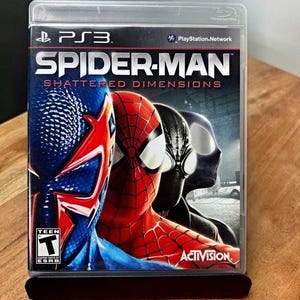 May include: A PlayStation 3 video game case for Spider-Man: Shattered Dimensions. The cover features three versions of the character in blue, red, and black suits. The title is printed in white and red text, with the ESRB Teen rating logo visible.