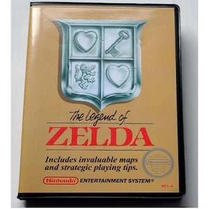 May include: A rectangular video game box for The Legend of Zelda on the Nintendo Entertainment System. The gold cover features a silver shield emblem with a heart, key, and lion, with the text Includes invaluable maps and strategic playing tips.