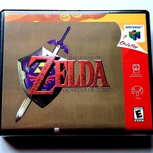 May include: A rectangular video game box for The Legend of Zelda: Ocarina of Time for Nintendo 64. The cover features a purple and silver shield with a sword, the game title in red text, the Nintendo 64 logo, and an ESRB Everyone rating symbol.