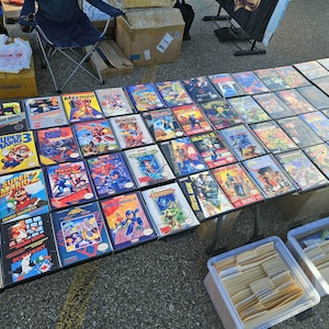 May include: A collection of vintage Nintendo Entertainment System video game boxes arranged on a table. The titles include Super Mario Bros, Mega Man, The Legend of Zelda, Final Fantasy, Metroid, and Metal Gear, featuring colorful cover art.