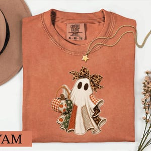 May include: A rust-colored t-shirt featuring a graphic of a ghost wearing a leopard print bow and holding a pumpkin-patterned basket. The ghost is decorated with various fabric-style patches in orange, green, and brown tones. The shirt has a Comfort Colors label.