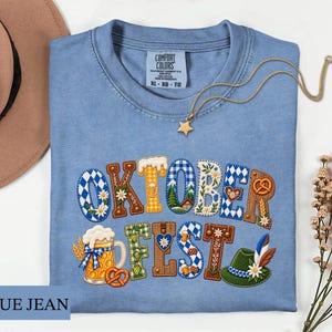 May include: A blue t-shirt featuring the text OKTOBERFEST in decorative letters filled with patterns of beer mugs, pretzels, Bavarian blue and white diamonds, flowers, and a green alpine hat. The shirt has a Comfort Colors label in the neck.