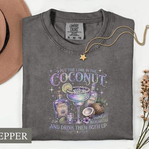 May include: A charcoal gray t-shirt featuring a graphic print of a margarita, tequila bottles, a coconut, and lime slices. The text reads: PUT THE LIME IN THE COCONUT AND DRINK THEM BOTH UP MIDNIGHT MARGARITAS. The shirt has a Comfort Colors label.