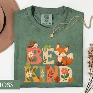 May include: A moss green short-sleeve t-shirt featuring a graphic print with the text BE KIND. The design includes a hedgehog, a chameleon, and a fox surrounded by flowers and autumn leaves. The shirt has a Comfort Colors brand label at the neck.