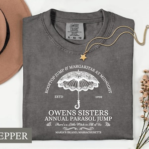May include: A charcoal gray t-shirt featuring a white graphic of a lace parasol. The text reads: ROOFTOP JUMP & MARGARITAS AT MIDNIGHT, ESTD 1998, OWENS SISTERS ANNUAL PARASOL JUMP, There's a Little Witch in All of Us, and MARIA'S ISLAND, MASSACHUSETTS.