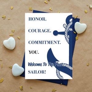 May include: A white greeting card with a dark blue anchor graphic wrapped in rope. The text reads: HONOR. COURAGE. COMMITMENT. YOU. Welcome To The Fleet SAILOR! The card is placed on a dark blue envelope, surrounded by small white heart-shaped stones.