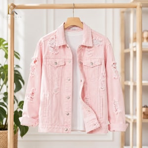 Puede incluir: Una chaqueta vaquera de color rosa claro con cuello clásico, mangas largas con puños doblados y cierre frontal de botones. La chaqueta presenta zonas desgastadas y deshilachadas en el pecho, los hombros y las mangas, e incluye dos bolsillos.