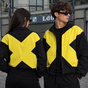 Puede incluir: Una chaqueta negra y amarilla con cremallera que presenta un llamativo patrón de bloques de color en forma de X en el pecho y la espalda. La chaqueta tiene cuello alto, mangas largas con puños acanalados y una base negra, sobre una camiseta negra.