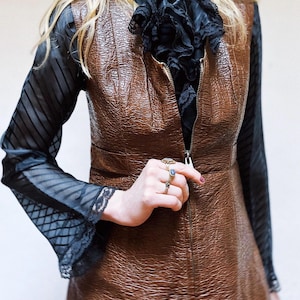 May include: A brown, textured dress with a zip fastening. The dress is layered over a black blouse with a ruffled collar and sheer sleeves with lace trim. Several rings are visible on the hand.