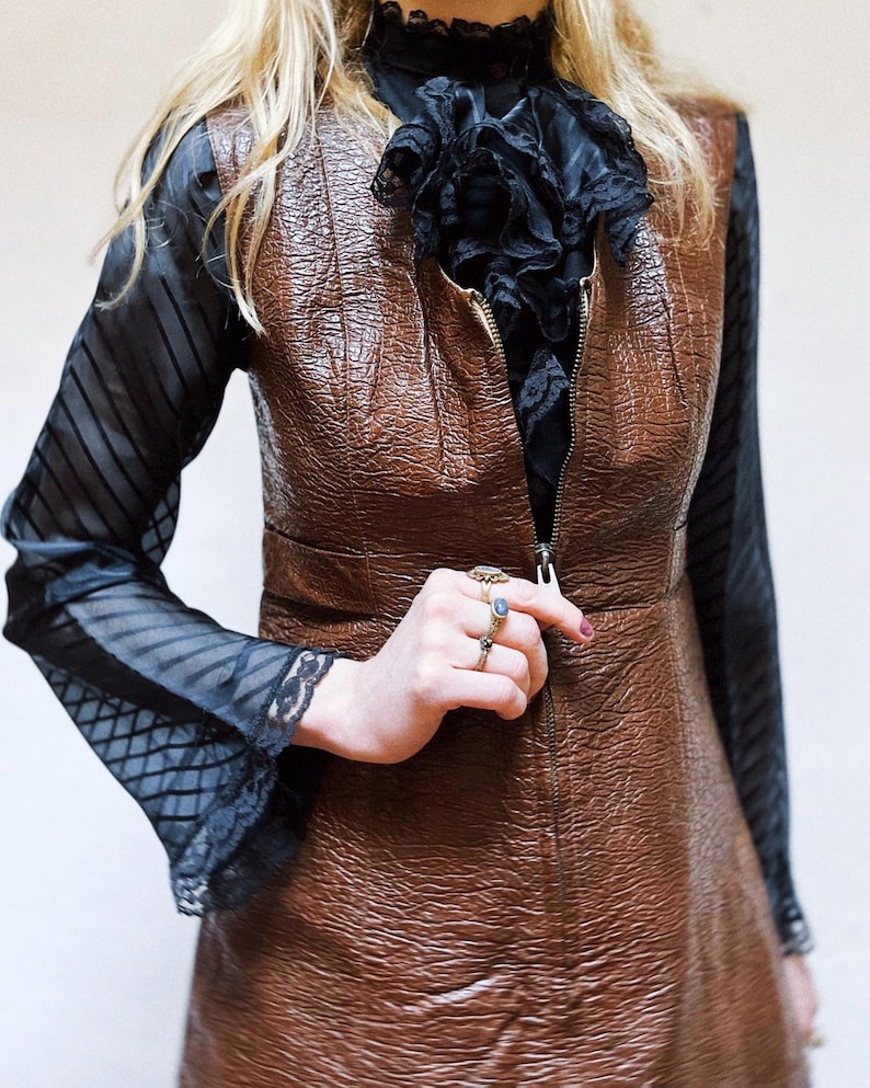 May include: A brown, textured dress with a zip fastening. The dress is layered over a black blouse with a ruffled collar and sheer sleeves with lace trim. Several rings are visible on the hand.