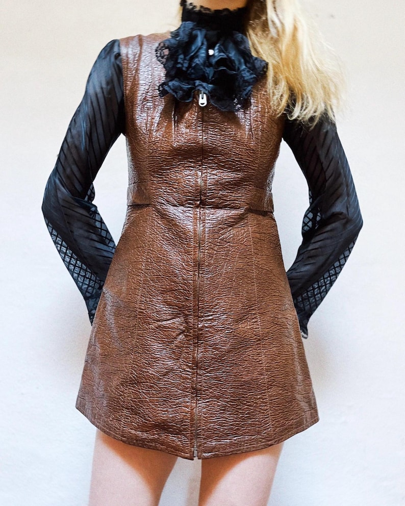 May include: A brown leather-look dress with a full-length zip down the front. The dress has a textured surface and a fitted silhouette. It is paired with a black sheer long-sleeve top with a patterned design on the sleeves and a black ruffled collar.