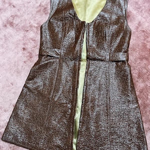 May include: A brown sleeveless vest with a textured finish and a V-neck. The vest features a full-length zip and a contrasting gold lining. A belt detail is present at the waist.