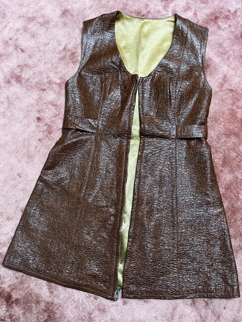 May include: A brown sleeveless vest with a textured finish and a V-neck. The vest features a full-length zip and a contrasting gold lining. A belt detail is present at the waist.