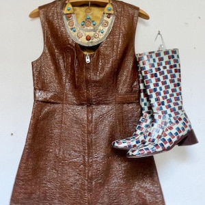 May include: A brown sleeveless dress with a zip, alongside patterned boots in white, red, and blue. A decorative necklace with turquoise and red accents hangs above the dress, creating a vintage aesthetic.