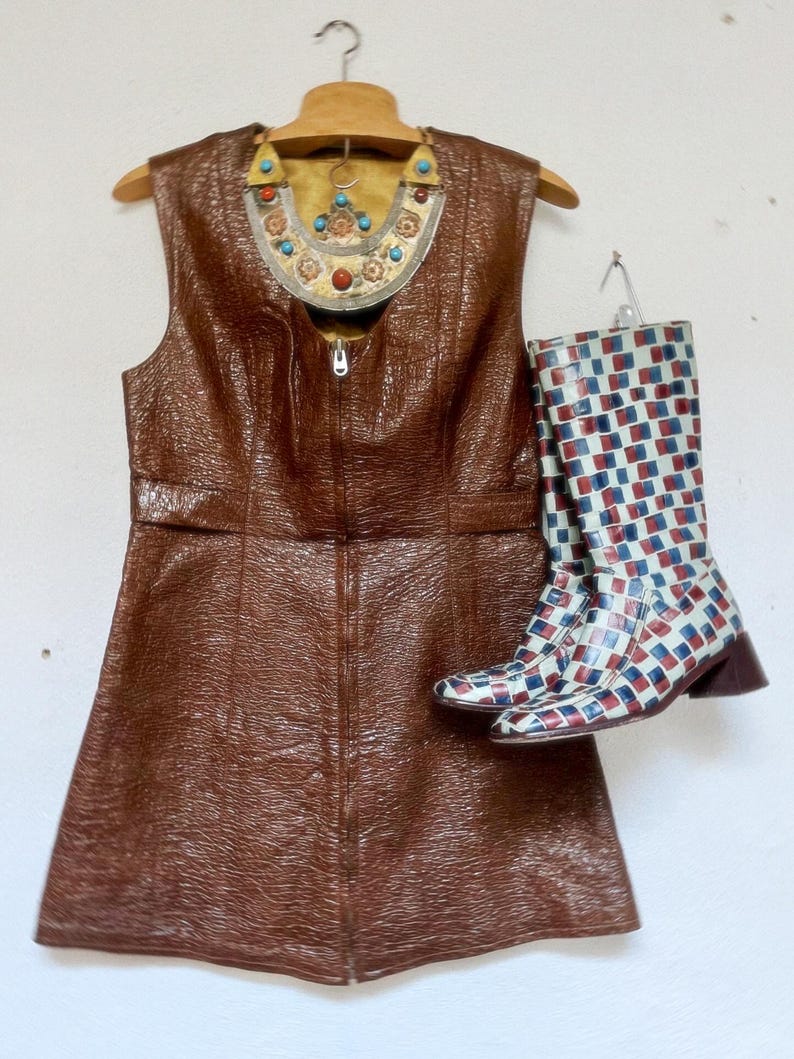 May include: A brown sleeveless dress with a zip, alongside patterned boots in white, red, and blue. A decorative necklace with turquoise and red accents hangs above the dress, creating a vintage aesthetic.