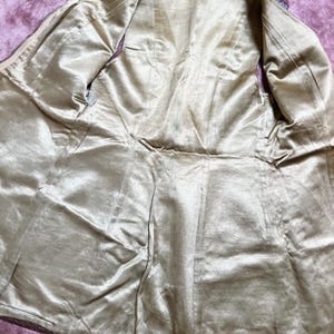 May include: An open, vintage-style jacket with a gold-coloured satin lining. The jacket's interior is visible, showcasing the lining's sheen and the tailored seams. The outer edges are trimmed with a dark, possibly leather, material.