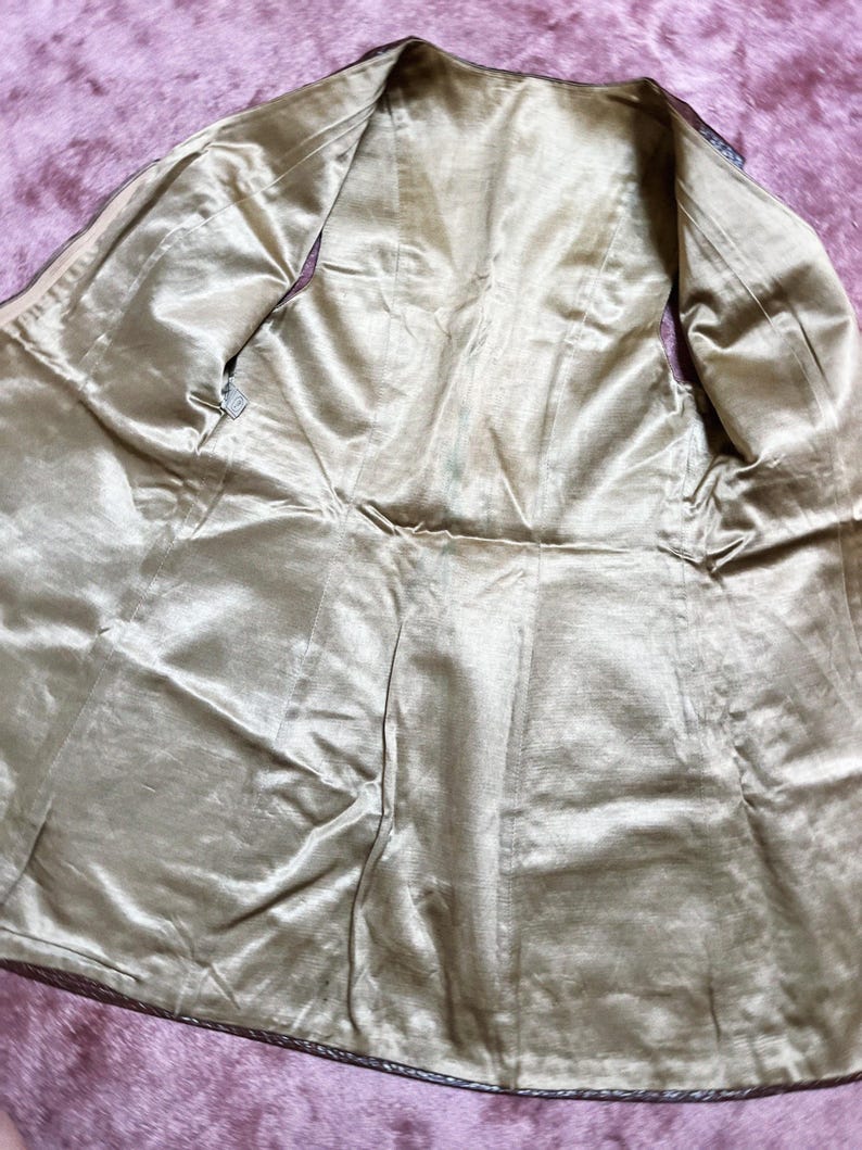 May include: An open, vintage-style jacket with a gold-coloured satin lining. The jacket's interior is visible, showcasing the lining's sheen and the tailored seams. The outer edges are trimmed with a dark, possibly leather, material.