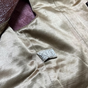 May include: Close-up of a brown leather jacket interior. The lining is a light gold satin material. A small tag with a geometric design is sewn into the lining. The jacket's exterior is a dark brown, textured leather.