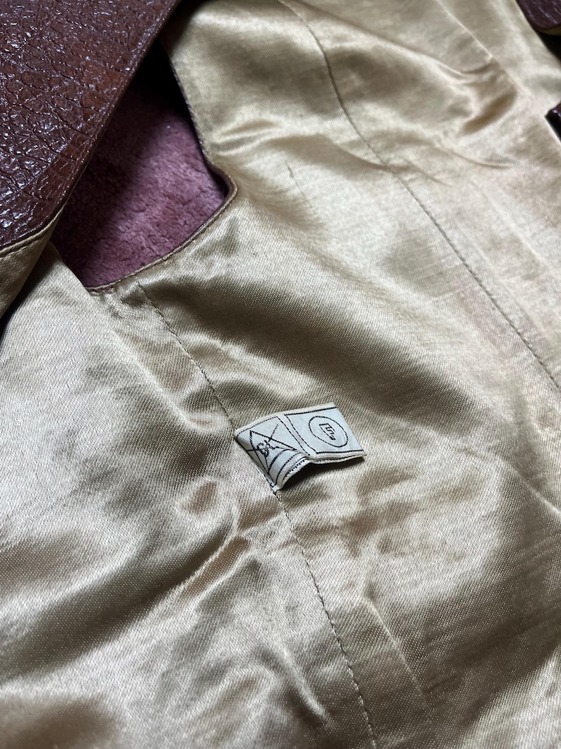 May include: Close-up of a brown leather jacket interior. The lining is a light gold satin material. A small tag with a geometric design is sewn into the lining. The jacket's exterior is a dark brown, textured leather.