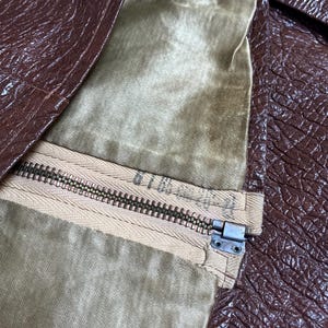 May include: Close-up of a brown leather jacket with a zipper. The jacket's interior lining is a light olive green colour. The zipper is a light tan colour with a metal zipper pull.