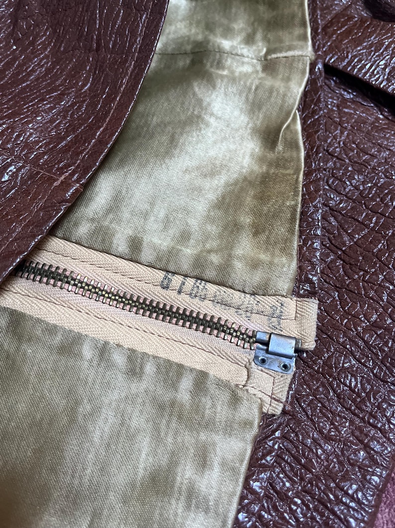 May include: Close-up of a brown leather jacket with a zipper. The jacket's interior lining is a light olive green colour. The zipper is a light tan colour with a metal zipper pull.