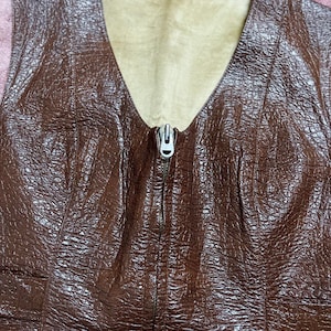 May include: A brown textured vest with a V-neck and a silver zip. The vest has a light brown lining and a detailed, embossed pattern. The design is a classic, vintage style.