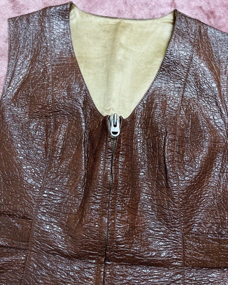 May include: A brown textured vest with a V-neck and a silver zip. The vest has a light brown lining and a detailed, embossed pattern. The design is a classic, vintage style.