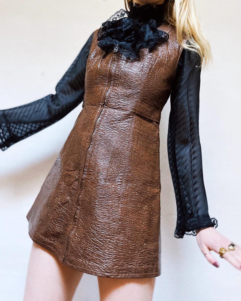 May include: A brown, textured, leather-look mini dress with a front zip. The dress is worn with a black sheer blouse with long sleeves and lace cuffs. A black ruffled collar adds a vintage touch to the outfit.