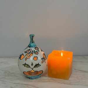Könnte beinhalten: Ein weißer Keramik-Ölbrenner in Tropfenform mit handgemalten orangefarbenen und grünen Blumenmotiven und einem blaugrünen Rand, neben einer quadratischen, durchscheinenden orangefarbenen Wachskerze mit brennendem Docht und warmem Licht.