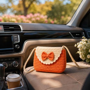 May include: A small, rectangular crochet crossbody bag featuring a vibrant orange main body and a cream-colored scalloped flap. The flap is adorned with a centered, textured orange bow. The bag includes a matching cream-colored crochet strap.