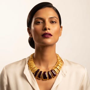 Puede incluir: Un collar llamativo que presenta una serie de segmentos de metal dorado texturizado dispuestos en estilo de cuello. La parte central delantera del collar muestra cinco inserciones rectangulares lisas de color burdeos profundo.