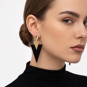 May include: A drop earring featuring a small gold-tone triangle at the top, a medium gold-tone triangle in the center, and a large black triangle at the base. The geometric shapes are stacked vertically to create a pointed, angular design.