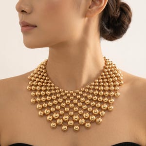 May include: A gold-toned statement necklace featuring a multi-layered, bib-style design composed of numerous polished, spherical beads connected in a cascading, net-like pattern that rests against the collarbone area with a high-shine metallic finish.