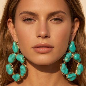 Puede incluir: Un par de pendientes grandes con piedras irregulares de color turquesa pulido, engastadas en un fino alambre de tono dorado. Las piedras están dispuestas en un diseño de aro circular abierto, creando un aspecto llamativo con tonos azul verdosos.