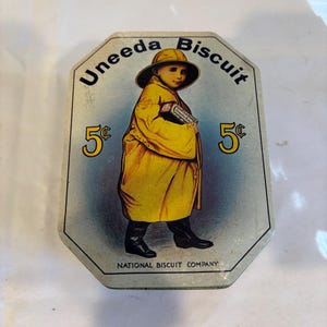 Puede incluir: Una lata de metal octogonal con una ilustración vintage de un niño con impermeable y sombrero amarillos sosteniendo una caja de Uneeda Biscuit. El texto dice Uneeda Biscuit, 5 centavos y National Biscuit Company en letras negras y amarillas.