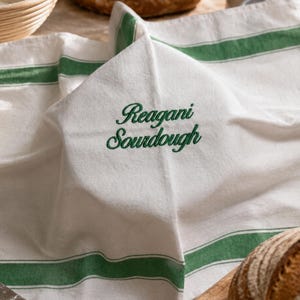 May include: A white kitchen towel featuring horizontal green stripes and the embroidered text Reagani Sourdough in a dark green cursive font. The fabric has a visible woven texture and is draped to show the embroidered lettering clearly.