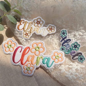 May include: Three embroidered fabric patches featuring the names Ethan, Clara, and Hazel in cursive script. Each patch is decorated with stitched flower motifs in various colours including orange, yellow, red, green, and blue on a white base.
