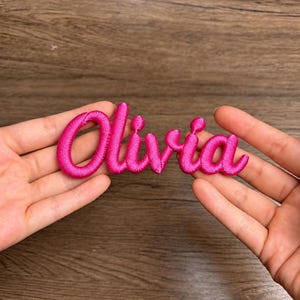 May include: A bright hot pink embroidered patch featuring the name Olivia in a cursive, rounded script font. The lettering has a raised, textured appearance with a consistent thread density throughout the entire name design.