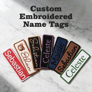 May include: Seven rectangular custom embroidered name tags arranged in a fan shape. The tags feature various colors including red, cream with a bear icon, black, purple, navy blue, brown, and white, each with stitched names like Sebastian, Ethan, Celeste, and Maisie.
