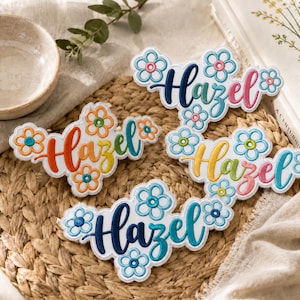 May include: Three embroidered patches featuring the name Hazel in cursive script, surrounded by stitched flower motifs. Each patch displays a unique colour gradient, including orange to yellow, blue to green, and dark blue to light blue tones.