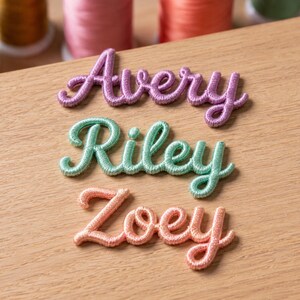 Puede incluir: Tres parches bordados con nombres en letra cursiva. El parche superior es morado y dice Avery, el del medio es verde menta y dice Riley, y el inferior es color melocotón y dice Zoey. Cada parche presenta costuras en relieve y texturizadas.