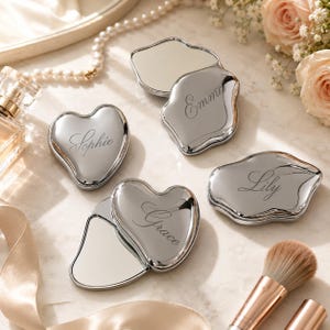 May include: Four silver-toned compact mirrors with irregular, organic shapes. Each mirror features a custom engraved name in a cursive script: Sophie, Emma, Lily, and Grace. The mirrors have a polished, reflective finish and are shown in both closed and open positions.
