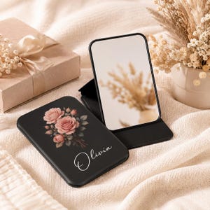 Puede incluir: Un espejo plegable rectangular de color negro con una ilustración impresa de rosas rosadas y el nombre Olivia en letra cursiva blanca. Un espejo está cerrado para mostrar el diseño floral y el nombre, mientras que el otro está abierto y refleja.