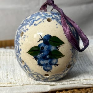 May include: A white ceramic ornament with a blue and white speckled glaze. The ornament features a hand-painted design of blueberries with green leaves. The ornament has a purple ribbon attached for hanging.