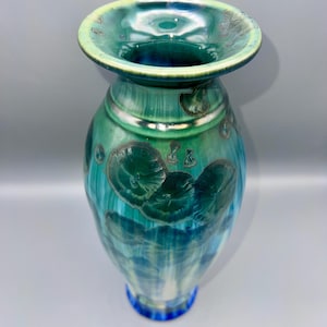 May include: A handcrafted ceramic vase with a wide, flared rim and a bulbous body. The vase features a blend of teal, turquoise, and blue hues with a glossy finish. Decorative circular patterns are visible on the surface.