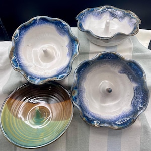 May include: Four ceramic apple bakers with a central stem for holding the apple. Three are white with blue rims, and one is brown and green. The bakers have a wavy edge and are on a striped cloth.